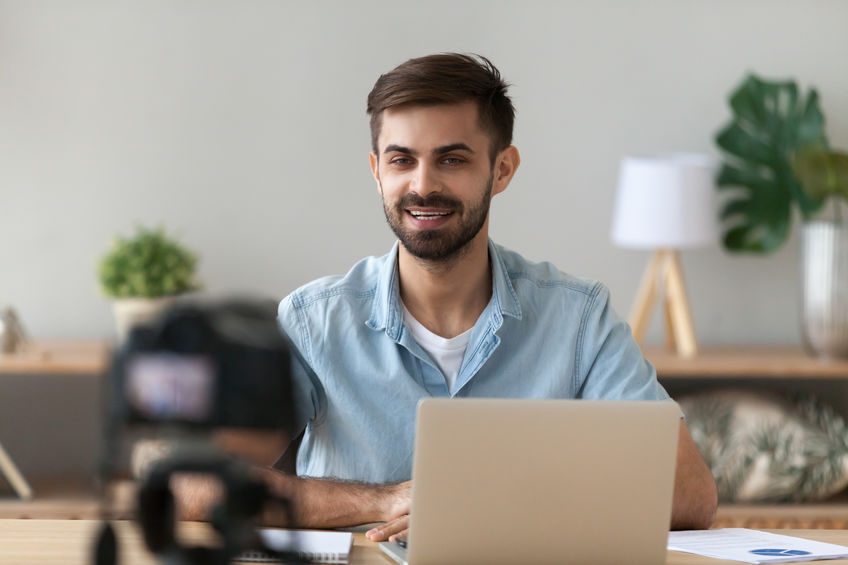 Young confident man speaker talking on digital camera recording vlog sitting at desk with laptop, male vlogger or business trainer speaking shooting blog filming reportage advertising video, coaching